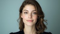Young Caucasian woman with natural makeup and wavy brown hair smiling confidently against turquoise background. Professional headshot style portrait showing genuine expression.