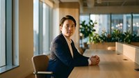 Young Asian businessman in navy suit sitting at conference table in modern office, hands clasped, serious expression, city view background.