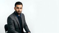 Adult male with dark hair and beard, Middle Eastern ethnicity, wearing charcoal blazer over dark shirt, seated against white background for corporate and professional use.