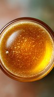 Golden honey in glass jar viewed from above showing crystalline texture and natural amber color with light reflections and bubbles creating organic pattern.