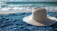 Straw sun hat on blue beach towel against blurred ocean waves and sparkling water background, summer vacation concept for travel and leisure.