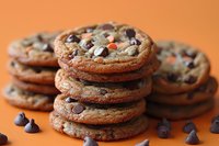 Stack of freshly baked chocolate chip cookies with crispy edges and soft centers on orange background, scattered chocolate chips create appetizing composition.