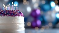 White frosted birthday cake with colorful sprinkles and glowing candles on blurred festive background with blue and purple bokeh lights and decorations.