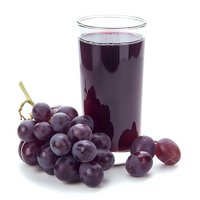Fresh purple grape juice in tall glass with ripe whole grapes scattered nearby on white background, healthy beverage and natural fruit composition.