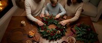 Caucasian family making Christmas wreath together at wooden table with pine cones, dried oranges, and red berries by cozy fireplace.