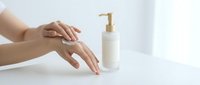 Woman applying hand cream from white pump bottle on clean surface for skincare routine and moisturizing treatment in bright minimalist setting.