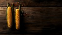 Fresh corn cobs with green leaves on dark rustic wooden background with copy space, dramatic lighting creates moody food photography atmosphere.