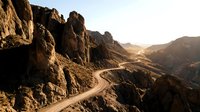 Winding mountain road through dramatic rocky canyon landscape with golden sunlight illuminating desert terrain and towering cliff formations, suitable for travel and adventure themes.