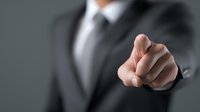 Businessman in formal suit pointing finger directly at viewer, conveying authority and leadership in professional corporate environment.