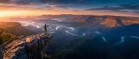 Hiker with arms raised celebrating success on mountain peak at sunrise with dramatic valley views and golden light illuminating rugged landscape below.