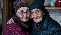 Two elderly women friends embracing and smiling warmly at camera, wearing winter clothing and glasses, showing authentic joy and companionship in their golden years.