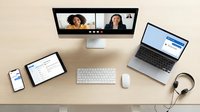 Modern remote work setup with video conference call on desktop monitor showing diverse team members during virtual meeting on wooden desk with laptop tablet and smartphone.