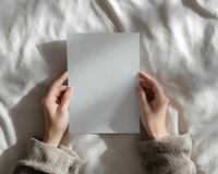 Female hands in cozy beige sweater holding blank white paper sheet against white textured fabric background with natural shadows and soft morning light.