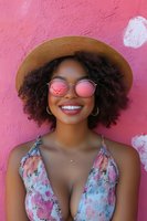 Young African American woman with natural curly hair wearing sunglasses and summer hat smiling against pink wall, reflecting carefree summer lifestyle and joy.