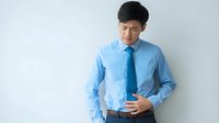 Asian businessman in blue shirt and tie experiencing stomach pain against light background. Concept for workplace stress, digestive issues, or health problems.