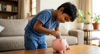 Young Asian boy in blue shirt saving money in pink piggy bank at home teaching financial literacy and responsibility for children's education.