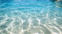 Tranquil turquoise water surface with sunlight creating rippling patterns and reflections, crystal clear shallow sea showing sandy bottom through transparent waves.