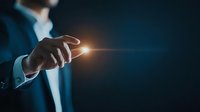Professional in dark suit pointing finger with glowing light effect against blue background, representing innovation, leadership and future technology solutions.