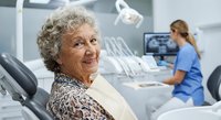 Happy elderly Caucasian woman smiling in modern dental office chair with female dentist working in background. Senior patient healthcare checkup visit.
