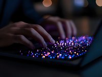 Hands typing on illuminated keyboard with vibrant purple and blue backlit keys creating magical glow effect in dark room, selective focus on glowing keys.