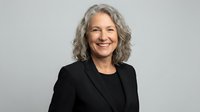 Professional mature Caucasian businesswoman with gray curly hair smiling confidently in black suit jacket against neutral background for corporate use.