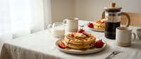 Fresh golden pancakes topped with strawberries and syrup on breakfast table with coffee press and milk pitcher in warm morning light kitchen setting.
