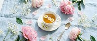 Herbal chamomile tea cup surrounded by pink peonies and white flowers on light blue linen background. Relaxing morning ritual flat lay composition.