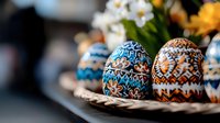Decorated Easter eggs with intricate folk patterns in blue, brown and white colors on woven plate with spring flowers in soft blurred background.