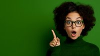 Young African American woman with curly afro hair and glasses showing surprised expression with raised finger against green background. Wearing green turtleneck sweater.