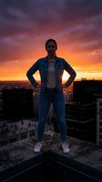 Confident young Caucasian woman in denim jacket stands on rooftop edge against dramatic fiery sunset sky over city skyline, hands on hips.