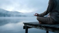 Person meditating in lotus position on wooden dock by misty mountain lake at dawn. Peaceful mindfulness and wellness concept for relaxation practices.