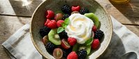 Fresh mixed berry fruit bowl with whipped cream, kiwi slices and mint garnish on rustic wooden table for healthy breakfast or dessert recipes.