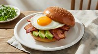 Gourmet bacon and egg burger with avocado slices on sesame seed bun served on white plate. Fresh breakfast sandwich with crispy bacon and sunny side up egg.