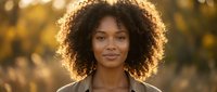 Young African American woman with curly hair smiling outdoors in warm golden sunlight during sunset or sunrise for lifestyle and portrait photography.