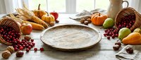 Autumn harvest arrangement with empty ceramic plate surrounded by fresh produce, wheat, nuts and berries on rustic wooden surface for seasonal food styling and recipe presentation.
