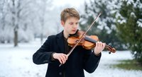 Young Caucasian male violinist performing outdoors in winter snow wearing formal black suit. Professional musician playing violin in snowy park landscape.