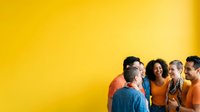 Group of diverse people wearing orange shirts standing together against bright yellow background, smiling and engaging in conversation for team building, community events, or corporate training materials.