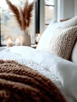 Cozy bedroom interior with knitted beige blanket, white bedding, decorative pampas grass, and glowing candles creating warm atmospheric lighting near window.