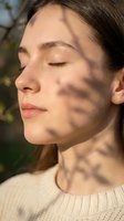 Young Caucasian woman with closed eyes enjoying warm sunlight with natural shadow patterns on her face in peaceful outdoor setting for wellness content.