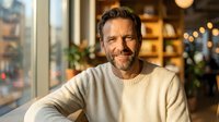 Handsome middle-aged Caucasian man with beard smiling warmly in cozy cafe setting. Professional portrait for lifestyle and business content.