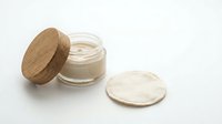 Natural organic skincare cream in glass jar with wooden lid and cotton pad on white background for beauty and wellness products.