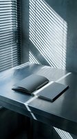 Minimalist workspace with open notebook and closed book on white desk, sunlight casting linear shadows through window blinds onto concrete wall, creating geometric patterns for design and editorial use.