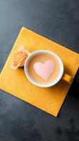 Cup of coffee with pink heart shape floating on top, placed on yellow napkin with peach rose, dark background. Perfect for Valentine's Day or romantic content.