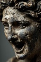 Ancient weathered stone sculpture face with dramatic expression and curly hair showing centuries of erosion and patina for historical art projects.