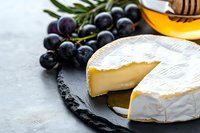 Soft ripened brie cheese wheel with cut piece on black slate board, fresh purple grapes and honey dipper, gourmet food styling on gray background.