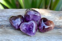 Purple amethyst heart shaped crystals on rustic wooden surface with soft natural lighting and green plant background, spiritual healing stones for meditation.