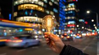 Hand holding glowing vintage light bulb against blurred city street at night with colorful bokeh lights and urban traffic creating innovation concept.