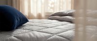 Cozy bedroom scene with soft quilted bedding, navy and gray pillows on white mattress, warm natural light filtering through sheer curtains, inviting sleep and relaxation.
