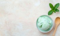 Mint ice cream scoop in ceramic bowl with fresh mint leaf and wooden spoon on light textured background, top view with copy space for menu design.