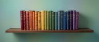 Rainbow colored books arranged in gradient order on a wooden shelf against a soft teal wall, creating a vibrant spectrum display.